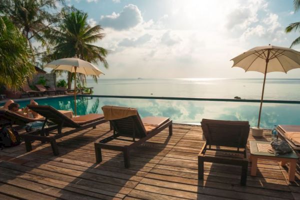 A serene setting with lounge chairs, umbrellas, and a pool overlooking the ocean, surrounded by palm trees under a partly cloudy sky.