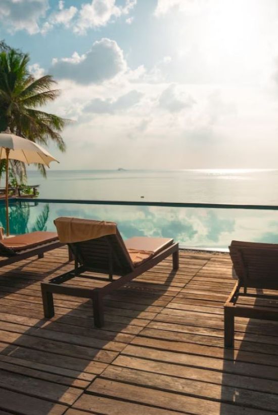 A serene poolside scene with lounge chairs, umbrellas, a calm infinity pool, and a stunning ocean view under a cloudy sky, glowing with sunlight.