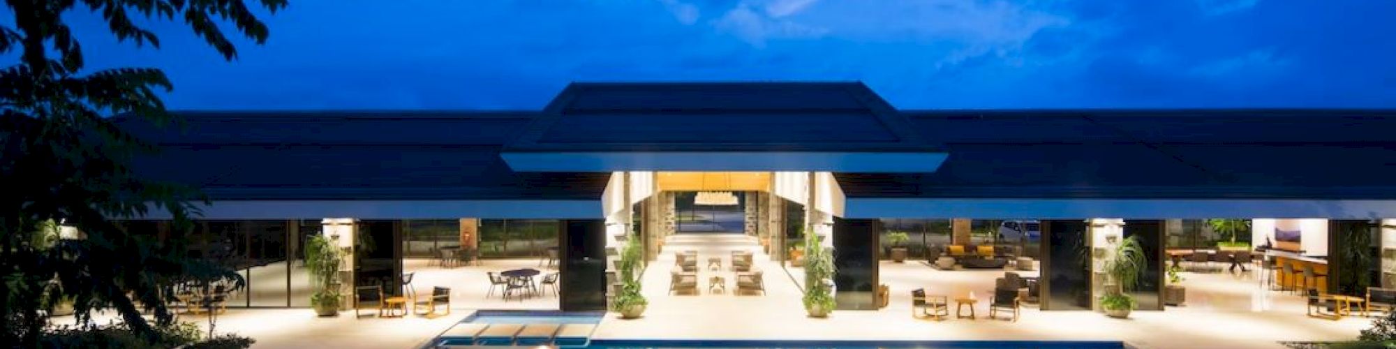 A luxurious outdoor pool area at dusk, featuring a modern building with open seating and dining spaces, illuminated with soft lighting at night.