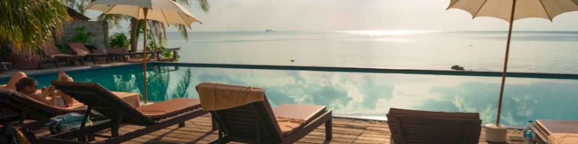 A serene poolside scene with lounge chairs and umbrellas facing a tranquil ocean, palm trees, and a bright sky in the background.