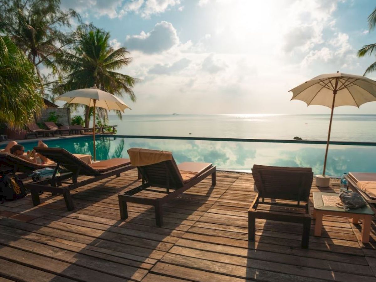 Poolside wooden deck with lounge chairs, umbrellas, and tropical scenery overlooking the ocean under a partly cloudy sky.