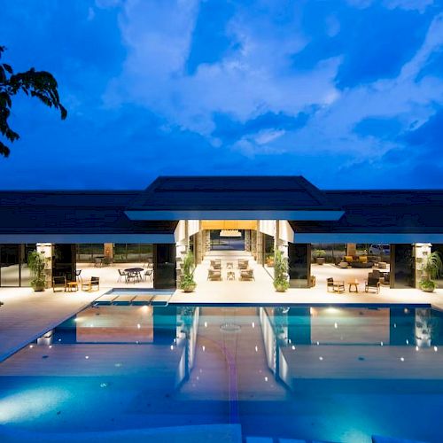 An illuminated swimming pool area with lounge chairs in front of a modern building at dusk, with a vibrant evening sky in the background.