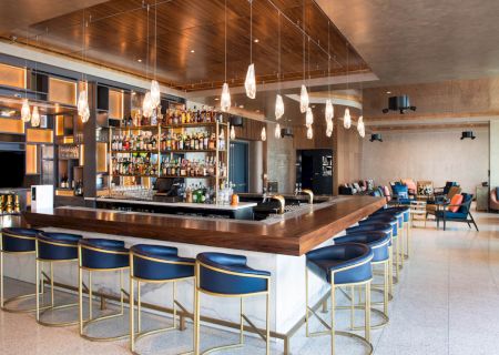 This image shows a modern bar with blue stools, a fully stocked bar, ambient lighting, and a seating area in the background.