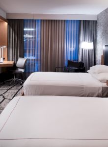 A modern hotel room with two neatly made beds, a wall-mounted TV, a desk with a chair, and contemporary decor featuring a patterned accent wall.