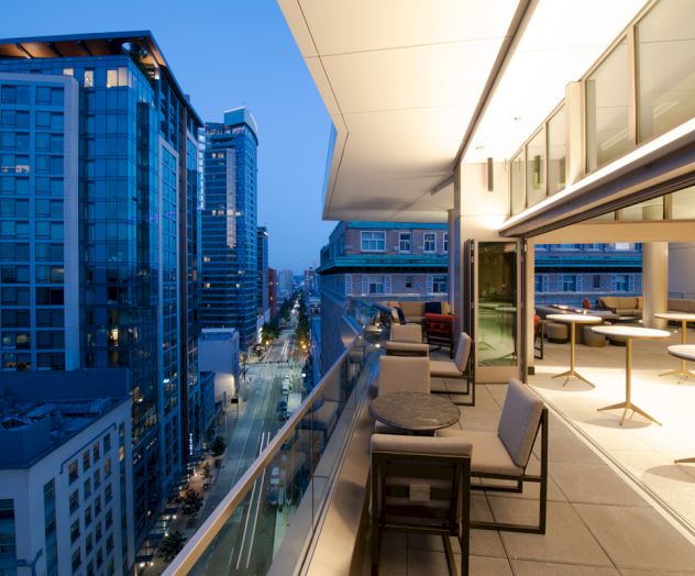 A modern cityscape view from a balcony at dusk, showing tall buildings, streetlights, and an outdoor seating area with tables and chairs.