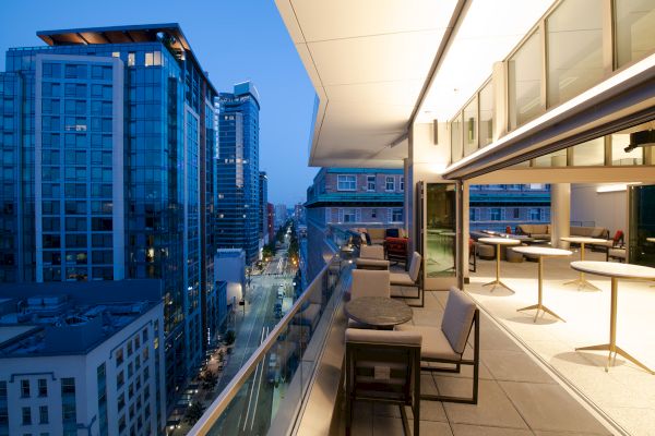 A modern cityscape at dusk with high-rise buildings and an outdoor balcony featuring tables, chairs, and a view of the illuminated street below.