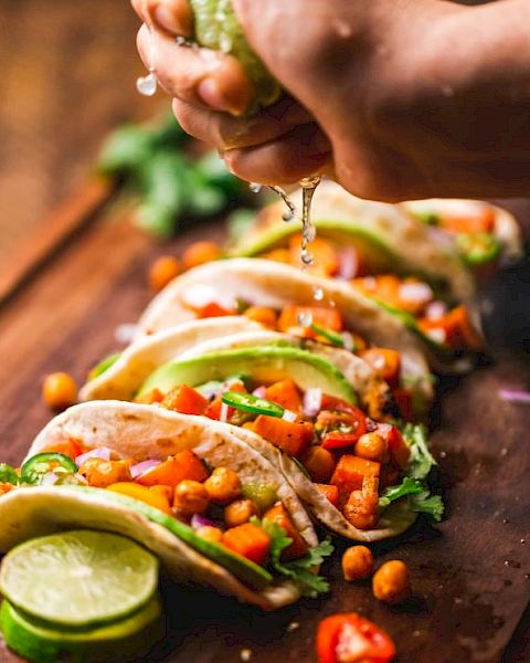 The image shows a hand squeezing lime juice over a row of tacos filled with chickpeas, diced tomatoes, avocado slices, and fresh herbs on a wooden board.