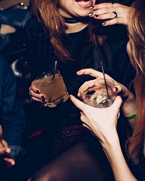 People socializing with drinks in a dimly lit setting, possibly a bar or a party, showing laughter and hand gestures.