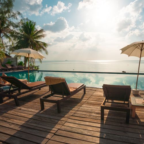 The image shows a serene beach setting with lounge chairs, umbrellas, and a pool deck facing a calm ocean under a partly cloudy sky.