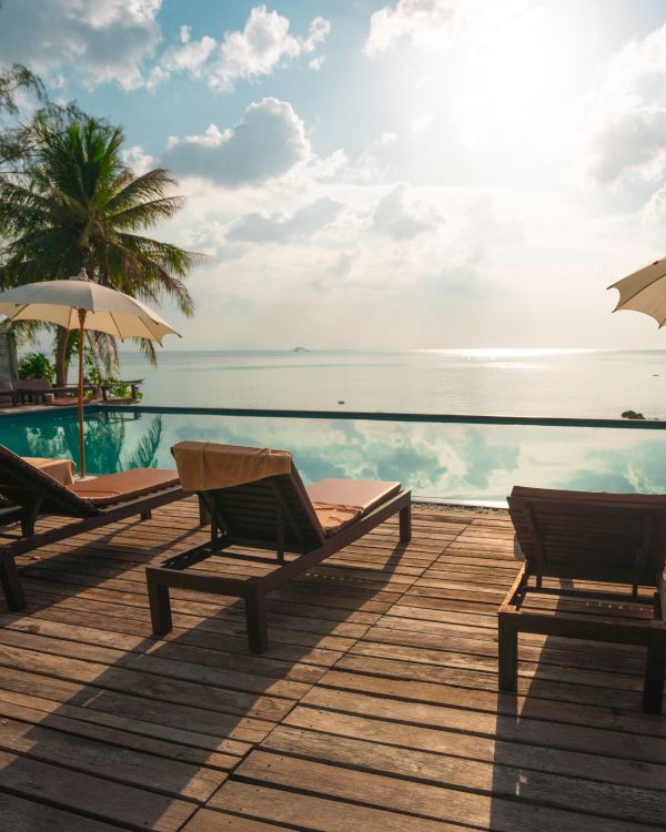 The image shows a serene beach setting with lounge chairs, umbrellas, and a pool deck facing a calm ocean under a partly cloudy sky.