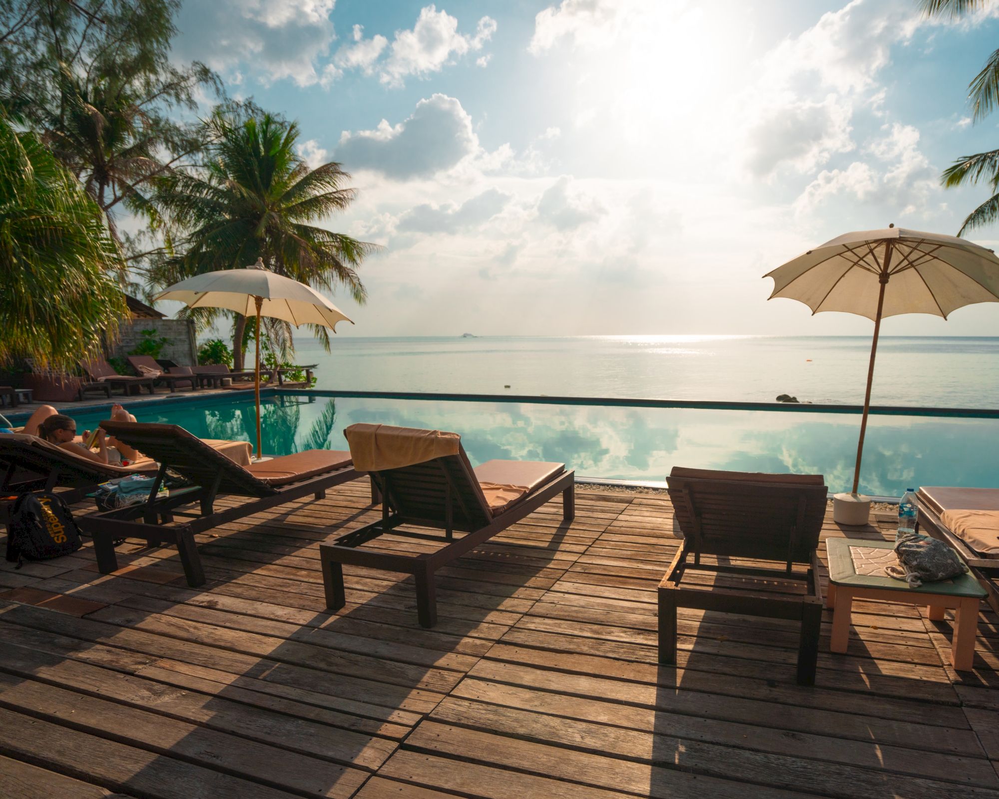 The image shows a serene seaside pool area with sun loungers, parasols, and wooden decking, overlooking the calm ocean amid palm trees and a sunny sky.