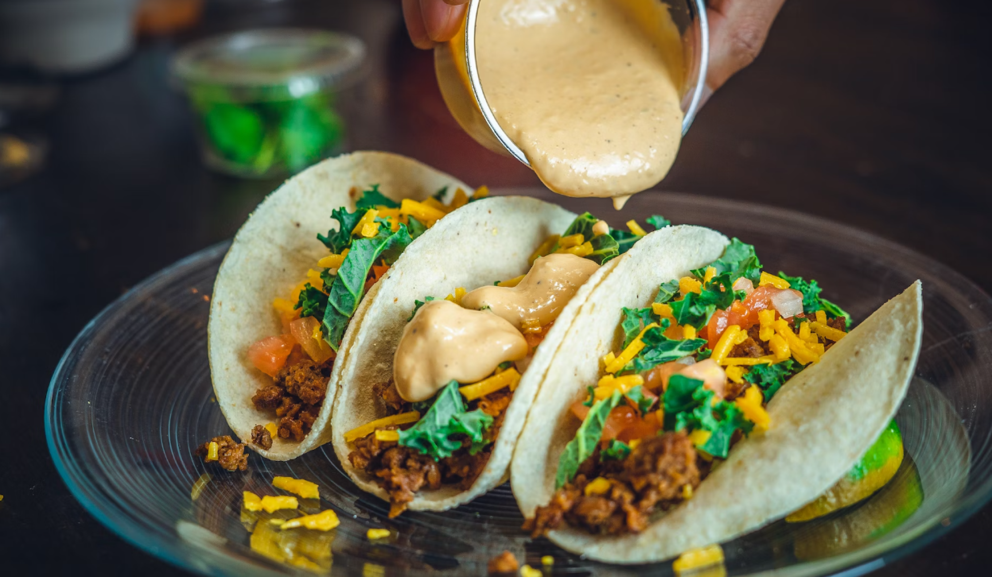 The image shows three tacos on a glass plate filled with meat, cheese, lettuce, and tomato, with someone pouring a creamy sauce on top.