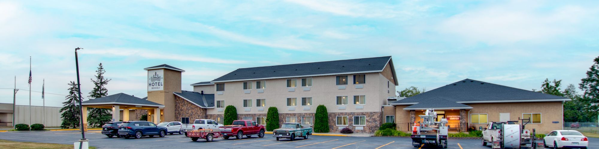 A building with a parking lot containing cars and a truck, set against a blue sky with clouds, is depicted.