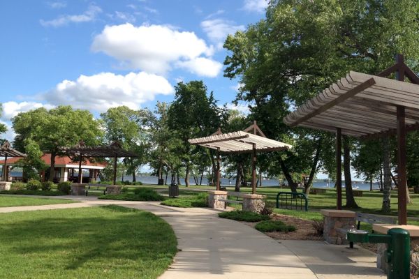 A sunny park with a walking path, green trees, and pavilions under a clear sky with fluffy clouds.