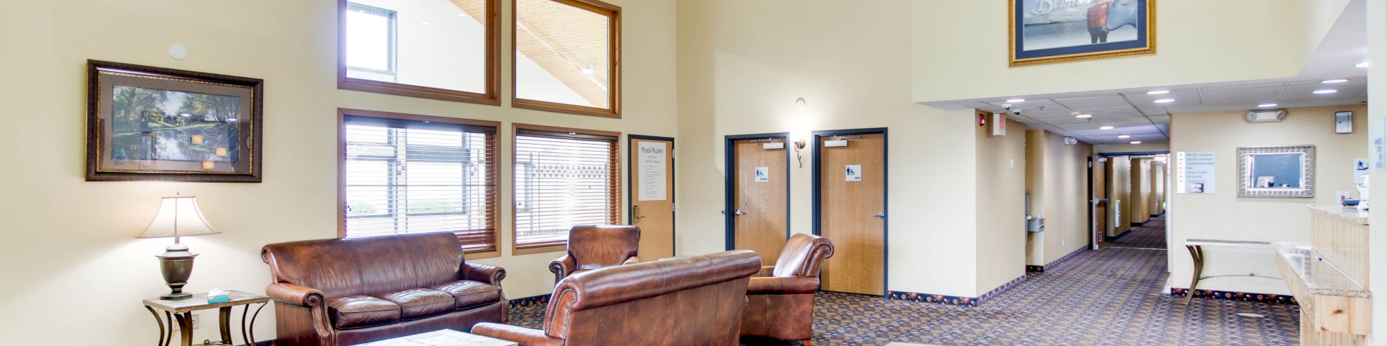 A spacious lobby with leather couches, tables, lamps, and framed pictures. The right-side hallway has several doors and a balcony above.