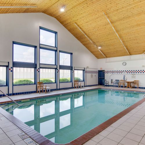 This image shows an indoor swimming pool with large windows, a high wooden ceiling, and tiled floors. The area includes poolside seating.