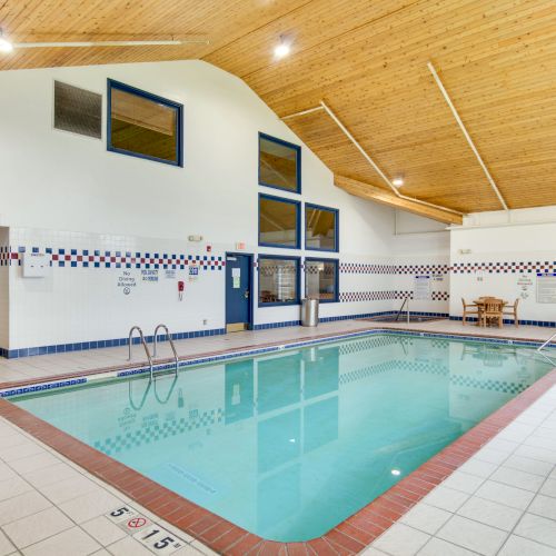 An indoor swimming pool with a wooden ceiling, surrounded by chairs and tables, with a 