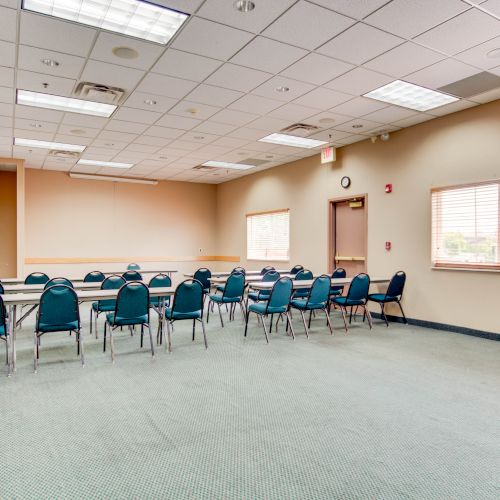 The image shows an empty, well-lit conference room with chairs arranged around tables in a U-shape on a green carpeted floor.