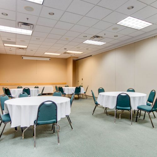 A conference room with round tables and green chairs, white tablecloths, and beige walls under a ceiling with fluorescent lighting.