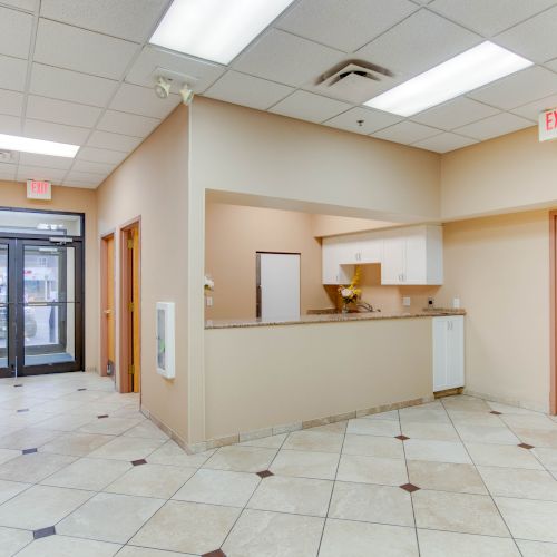 A clean tiled hallway with beige walls, a reception counter, exit doors, a door on the right, and a dolly cart against the wall.