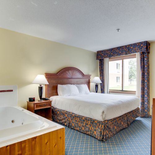 The image shows a hotel room with a king-sized bed, nightstands, lamps, a framed picture, a window, a TV cabinet, and a jacuzzi tub, in a cozy setting.