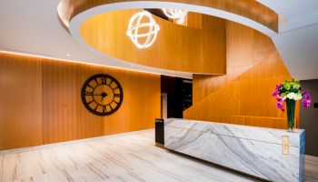 The image shows a modern lobby with a curved wooden ceiling feature, a large wall clock, a marble reception desk, and a vase of flowers.