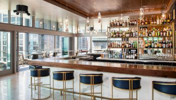 The image shows a modern bar with a sleek design, featuring a row of bar stools, a well-stocked shelf of liquor, and a view of the city through large windows.