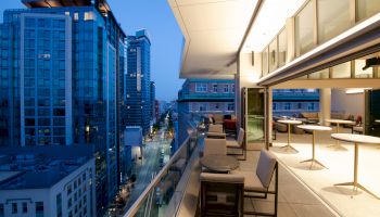 This image shows a balcony view from a modern high-rise building, overlooking a cityscape with illuminated skyscrapers and a street below at dusk.