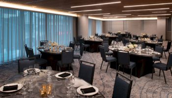 The image shows an elegantly set dining hall with round tables covered in black tablecloths, neatly arranged chairs, floral centerpieces, and glassware.