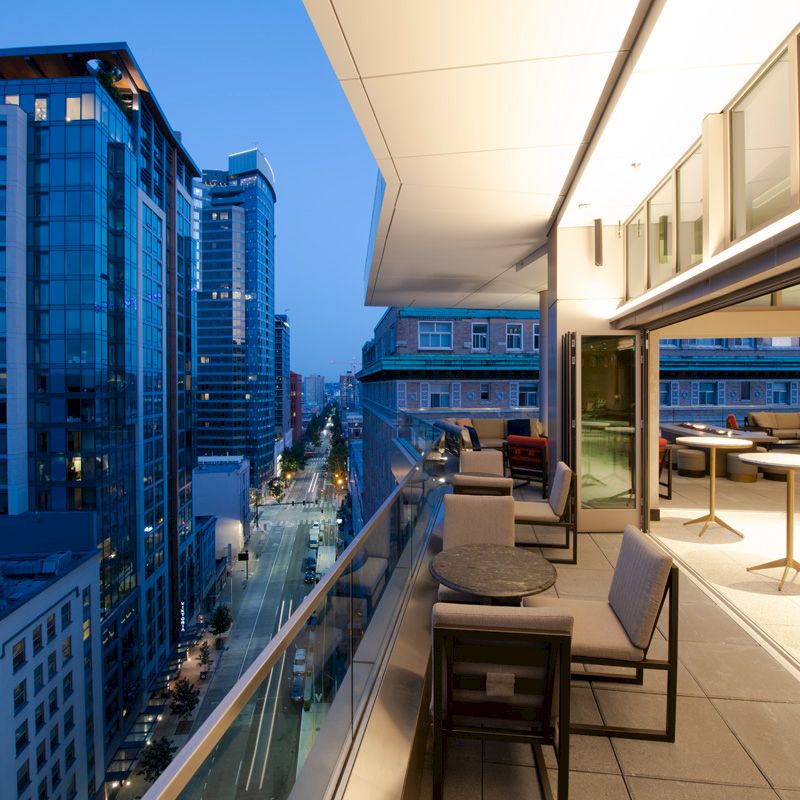 A view of a modern cityscape at dusk from an outdoor terrace with seating and tables, adjacent to a building with large windows and lights.