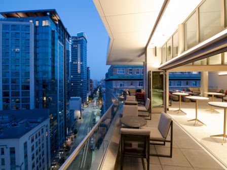 A modern rooftop patio with tables and seating overlooking an urban cityscape at twilight, with tall buildings lining the street below.