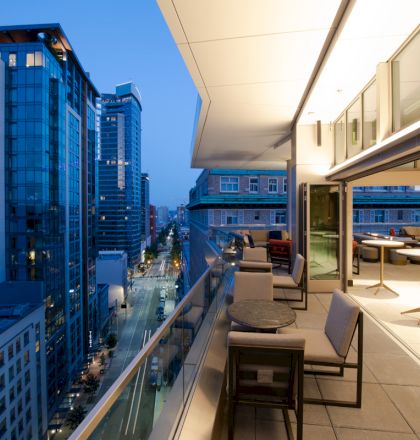 The image shows a modern balcony at dusk with seating and tables, overlooking a city street lined with tall buildings and glowing lights.