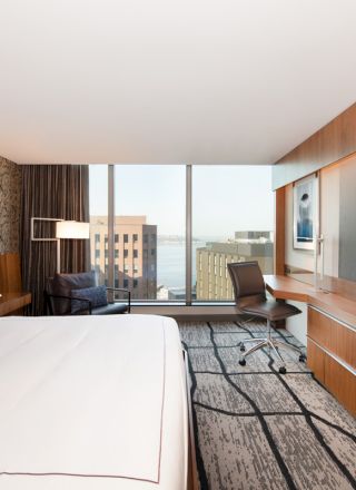 This image shows a modern hotel room with a large bed, wall-mounted TV, desk, and a view of tall buildings through the window.