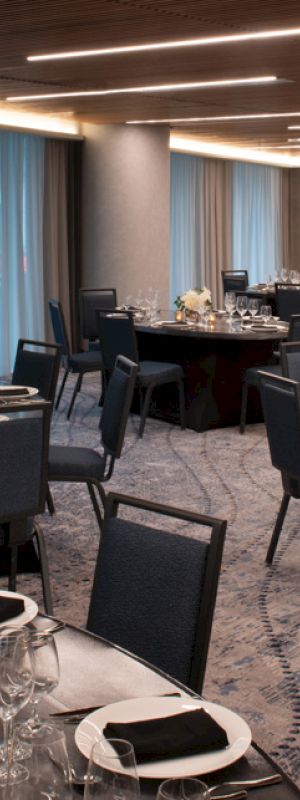 The image shows an elegant dining hall set up for an event with round tables, black chairs, place settings, and floral centerpieces on each table.