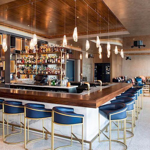 A modern bar with a wooden counter, blue bar stools, and hanging lights. Shelves with liquor bottles are behind the counter, with seating areas around.