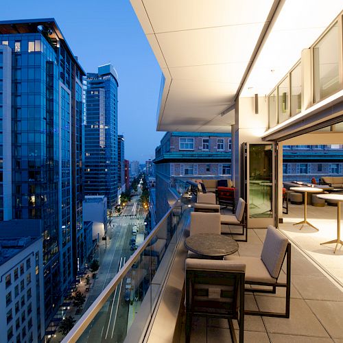 High-rise cityscape view with modern buildings at dusk, and an outdoor terrace featuring tables and chairs on the right side of the image.